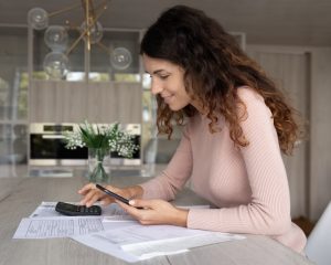 Smiling millennial Latin female manage household budget expenditures pay online on cellphone. Happy young Hispanic woman count on calculator machine make payment on smartphone. Finances concept.