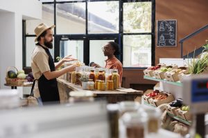 Black woman receiving product recommendations from caucasian vendor at a zero waste supermarket. In a nearby eco friendly shop, male seller assists african american customer.
